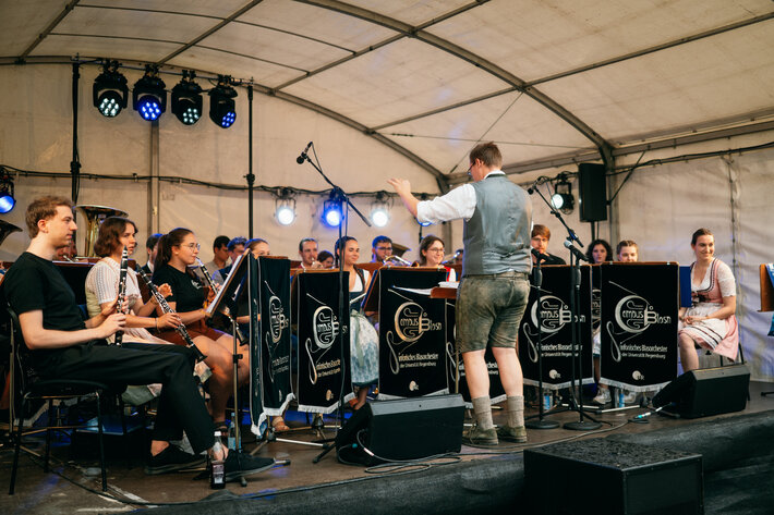 Auftritt der CampusBlosn: junge Musikerinnen und Musiker mit Blasinstrumenten sitzen auf der Bühne und spielen ein Musikstück. Sie werden von einem jungen Mann in Tracht dirigiert. Sie selbst tragen teilweise ebenfalls Tracht. (öffnet Vergrößerung des Bildes)