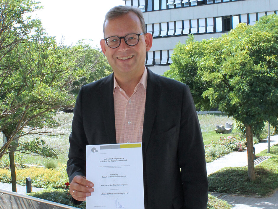 Prof. Kingreen steht lächelnd mit der Urkunde in Händen vor dem Rosengarten neben dem Hörsaalgebäude der Juristischen Fakultät. (öffnet Vergrößerung des Bildes)