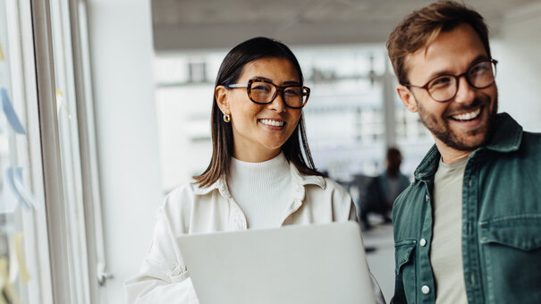 Eine Frau mit Brill und einem Laptop in der Hand steht lächelnd neben einem Mann mit Brille und freundlichem Gesichtsausdruck