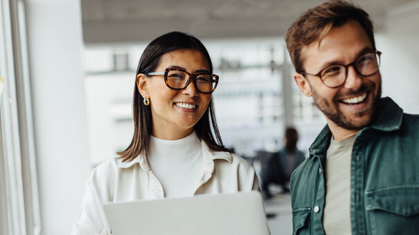 Eine Frau mit Brill und einem Laptop in der Hand steht lächelnd neben einem Mann mit Brille und freundlichem Gesichtsausdruck
