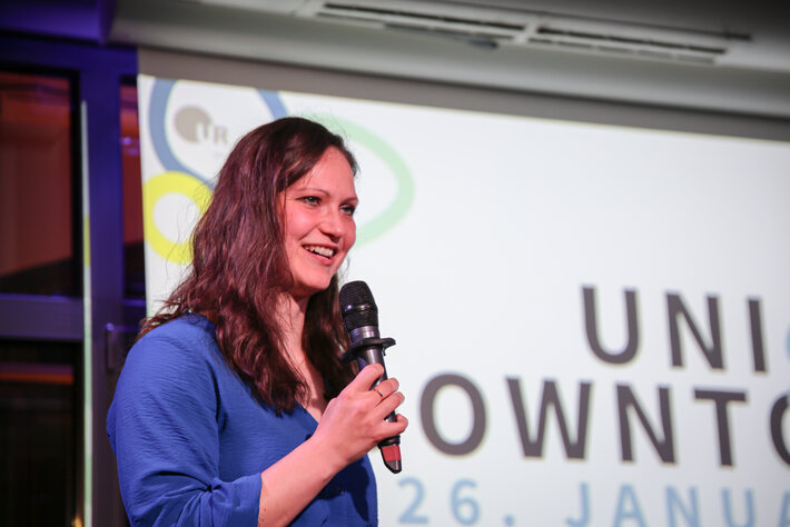 Eine Person steht vor einer großen, hellen Projektionsfläche und hält ein Mikrofon in der Hand. Auf der Leinwand hinter ihr sind Teile eines Veranstaltungslogos und Schriftzugs zu erkennen. Die Person trägt ein blaues Oberteil und scheint auf einer Bühne oder bei einer Präsentation zu sprechen. (öffnet Vergrößerung des Bildes)