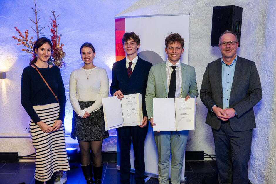 Zwei Frauen und drei Männer stehen nebeneinander. Die beiden jüngeren Männer halten Urkunden in die Kamera.