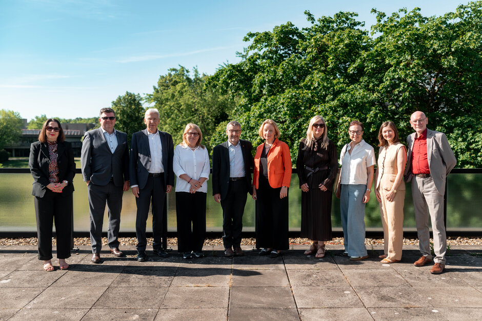 Elf Personen stehen nebeneinander auf einer sonnigen Terrasse vor einem Geländer. Im Hintergrund sind große grüne Bäume und ein klarer blauer Himmel zu sehen. Die Personen tragen überwiegend Business- oder Business-Casual-Kleidung in dunklen und hellen Farben und blicken in Richtung Kamera. Die Aufnahme zeigt eine Gruppenaufstellung im Freien bei Tageslicht. (öffnet Vergrößerung des Bildes)
