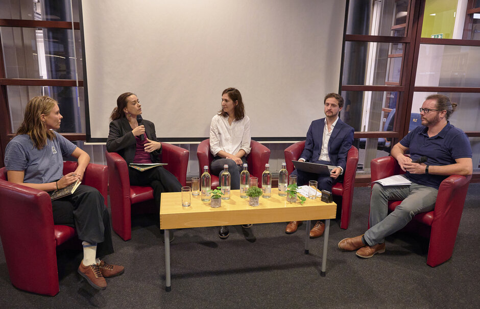 Fünf Personen mit Mikrofonen sitzend an Tisch, sprechen miteinander