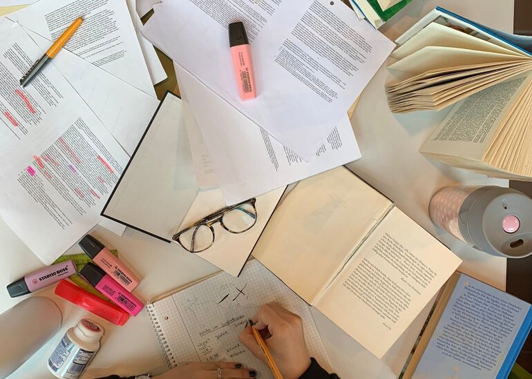 Person, die vor vielen Zetteln und Büchern sitzt und lernt.