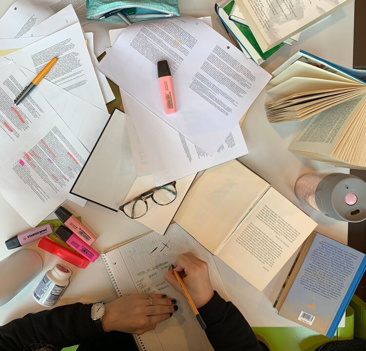 Person, die vor vielen Zetteln und Büchern sitzt und lernt.