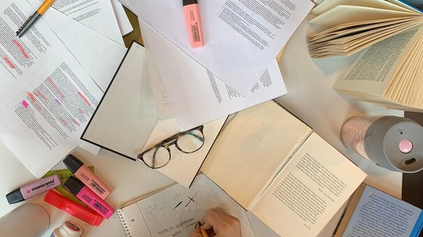 Person, die vor vielen Zetteln und Büchern sitzt und lernt.