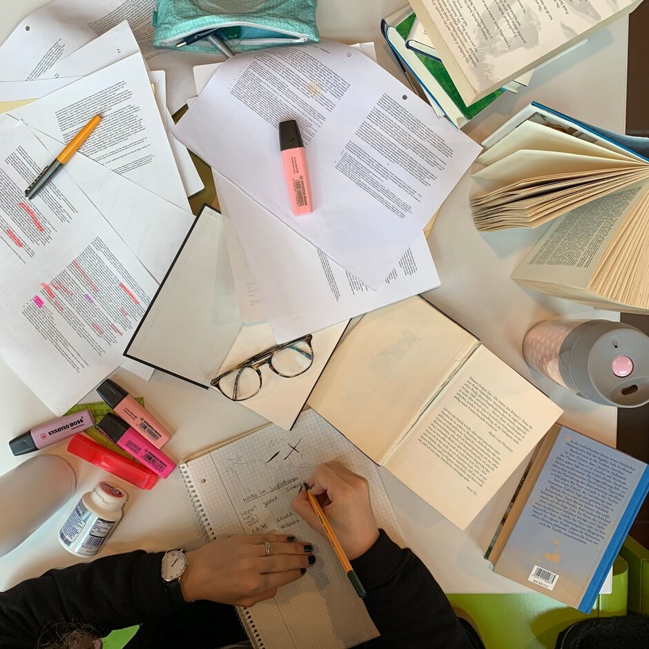 Person, die vor vielen Zetteln und Büchern sitzt und lernt.