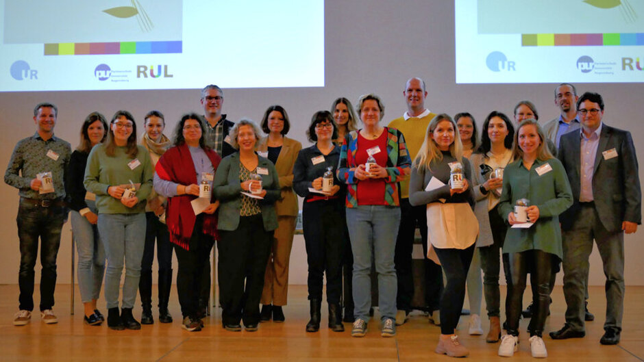 Gruppenbild mit mehreren Personen, die alle Namensschilder tragen. Sie stehen in einer losen Reihe auf einer Bühne und halten Tassen mit der Aufschrift RUL in den Händen.