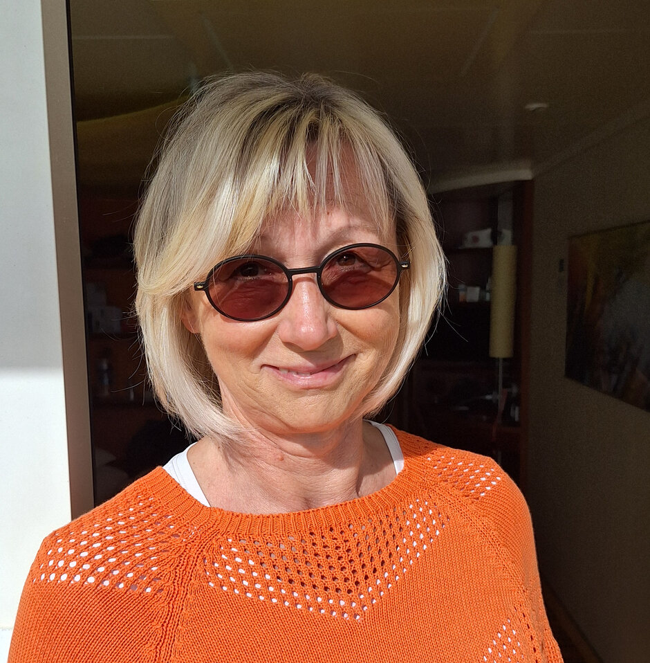 Eine blonde Frau mit Sonnenbrille lächelt den Betrachter an. Sie trägt einen orangen Pullover.