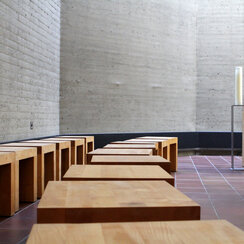 Ein schlichter Raum mit Sichtbeton-Wand und gefließtem Boden. An der Wand lässt sich ein schlichtes Metallkreuz erahnen, eine hohe weiße Kerze steht auf einem Kerzenhalter, ein Holztisch ist mit einem Blumenstrauße geschmückt. Entlang der Wand stehen zwei Reihen schlichter Holzhocker., Schlichte Holzhocker stehen in zwei lockeren Reihen und führen 