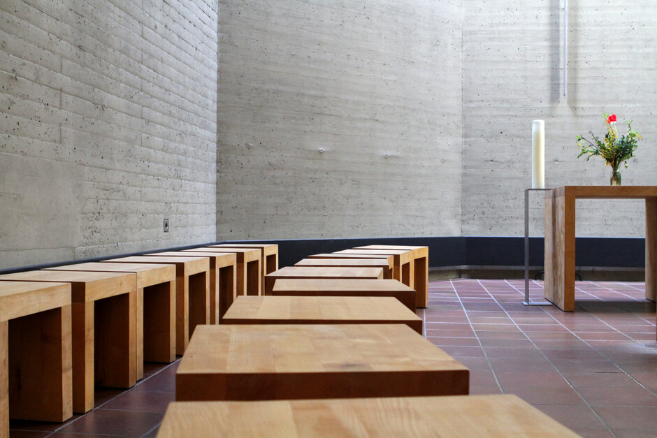 Ein schlichter Raum mit Sichtbeton-Wand und gefließtem Boden. An der Wand lässt sich ein schlichtes Metallkreuz erahnen, eine hohe weiße Kerze steht auf einem Kerzenhalter, ein Holztisch ist mit einem Blumenstrauße geschmückt. Entlang der Wand stehen zwei Reihen schlichter Holzhocker., Schlichte Holzhocker stehen in zwei lockeren Reihen und führen 