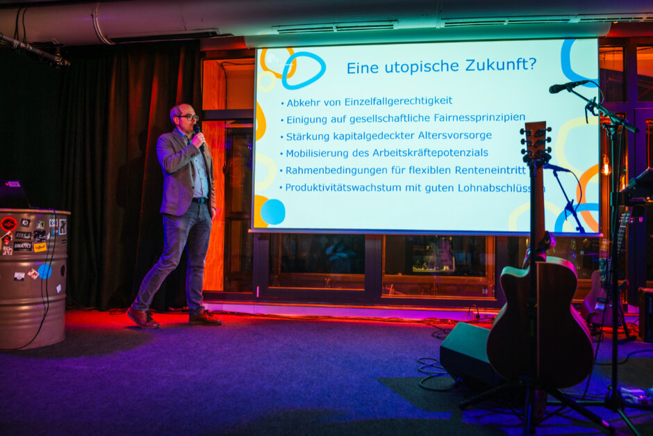 Ein Mann steht mit einem Mikrophon in der Hand auf einer Bühn und erklärt seine Präsentation.