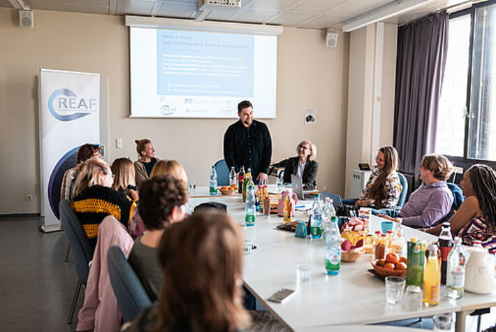 Ein Konferenzraum mit einem breiten Tisch, auf dem Getränke und Obst stehen. Um den Tisch herum sitzen mehrere Personen, die einem Mann zuhören, der am kurzen Ende des Tisches steht und spricht. (öffnet Vergrößerung des Bildes)