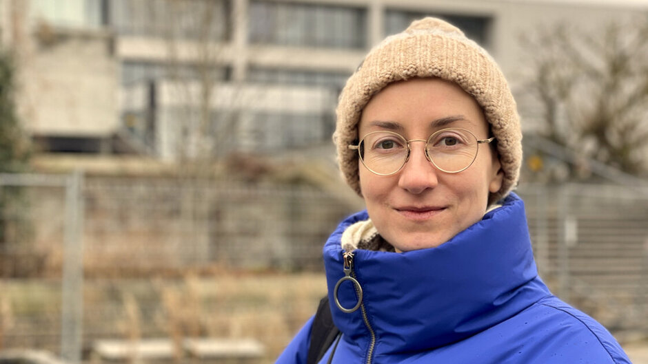 Eine junge Frau mit Brille, Wollmütze und blauer Winterjacke steht vor einem Hochschulgebäude