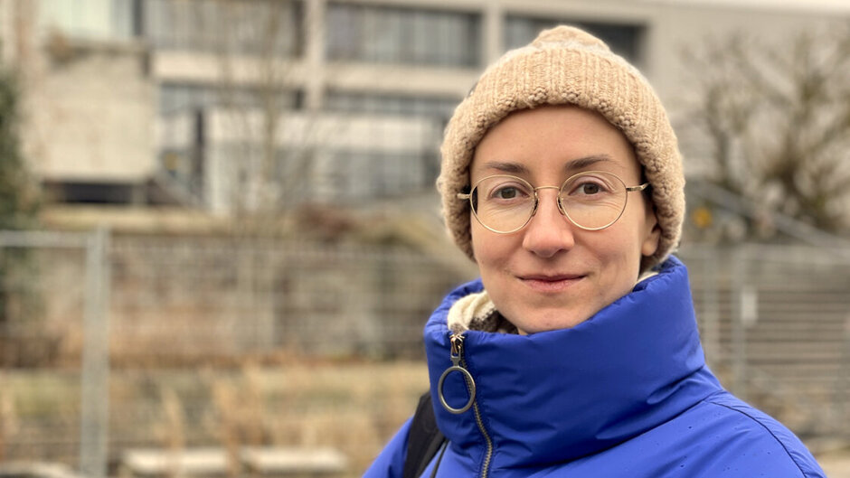 Eine junge Frau mit Brille, Wollmütze und blauer Winterjacke steht vor einem Hochschulgebäude