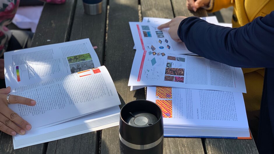 Auf einem Tisch sind Bücher aufgeschlagen, ein Kaffeebecher steht daneben