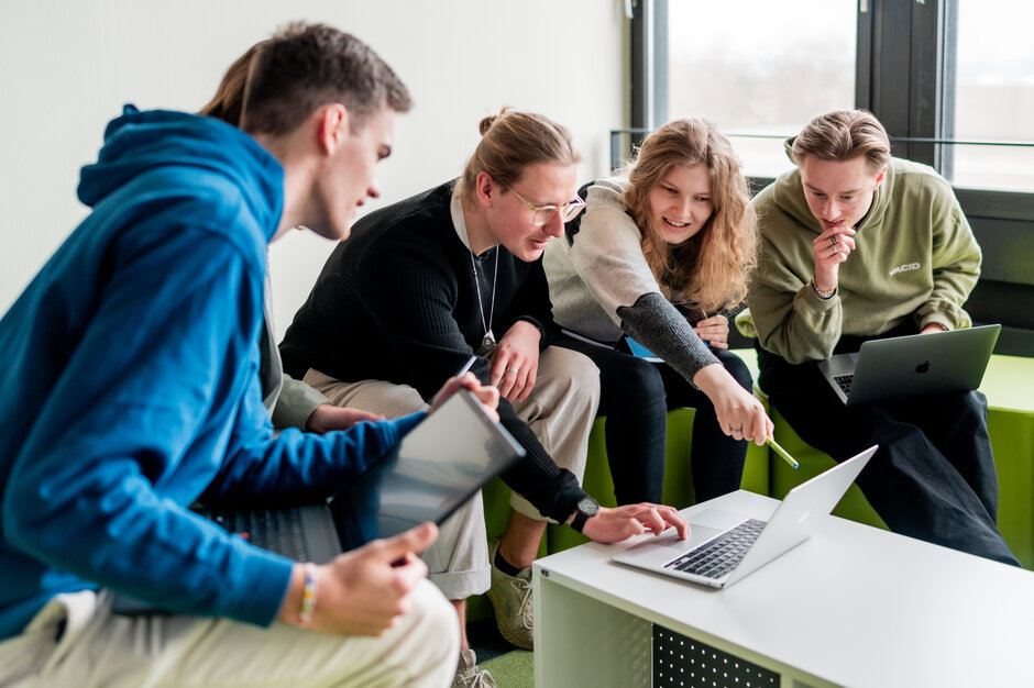 Studierende sitzen gemeinsam um einen Laptop und diskutieren über das auf dem Bildschirm gezeigte.