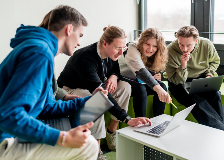 Studierende sitzen gemeinsam um einen Laptop und diskutieren über das auf dem Bildschirm gezeigte.