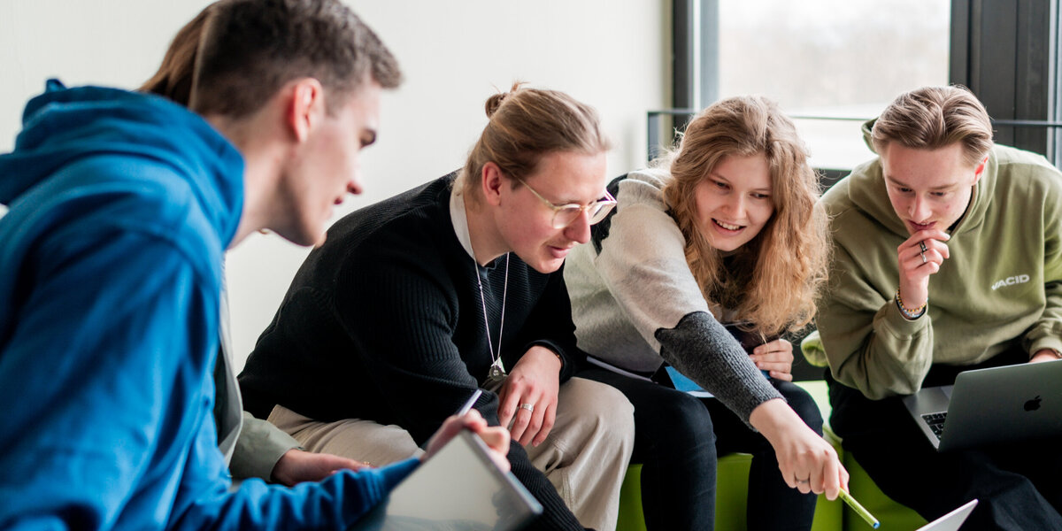 Studierende sitzen gemeinsam um einen Laptop und diskutieren über das auf dem Bildschirm gezeigte.