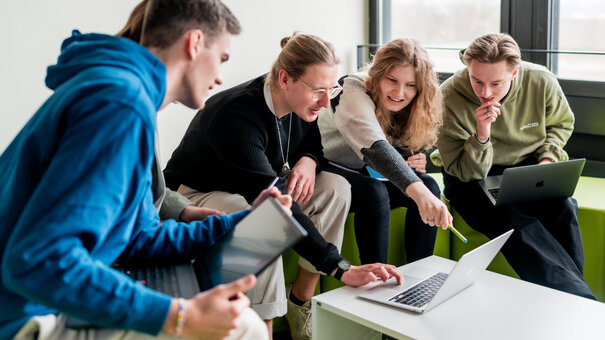Studierende sitzen gemeinsam um einen Laptop und diskutieren über das auf dem Bildschirm gezeigte.
