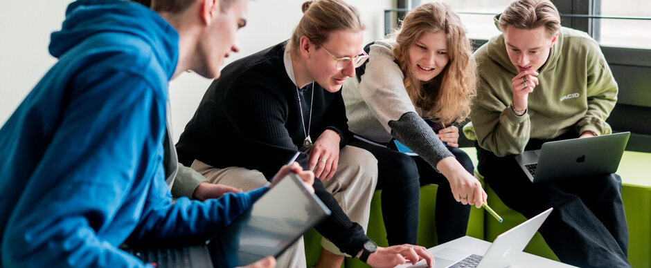 Studierende sitzen gemeinsam um einen Laptop und diskutieren über das auf dem Bildschirm gezeigte.