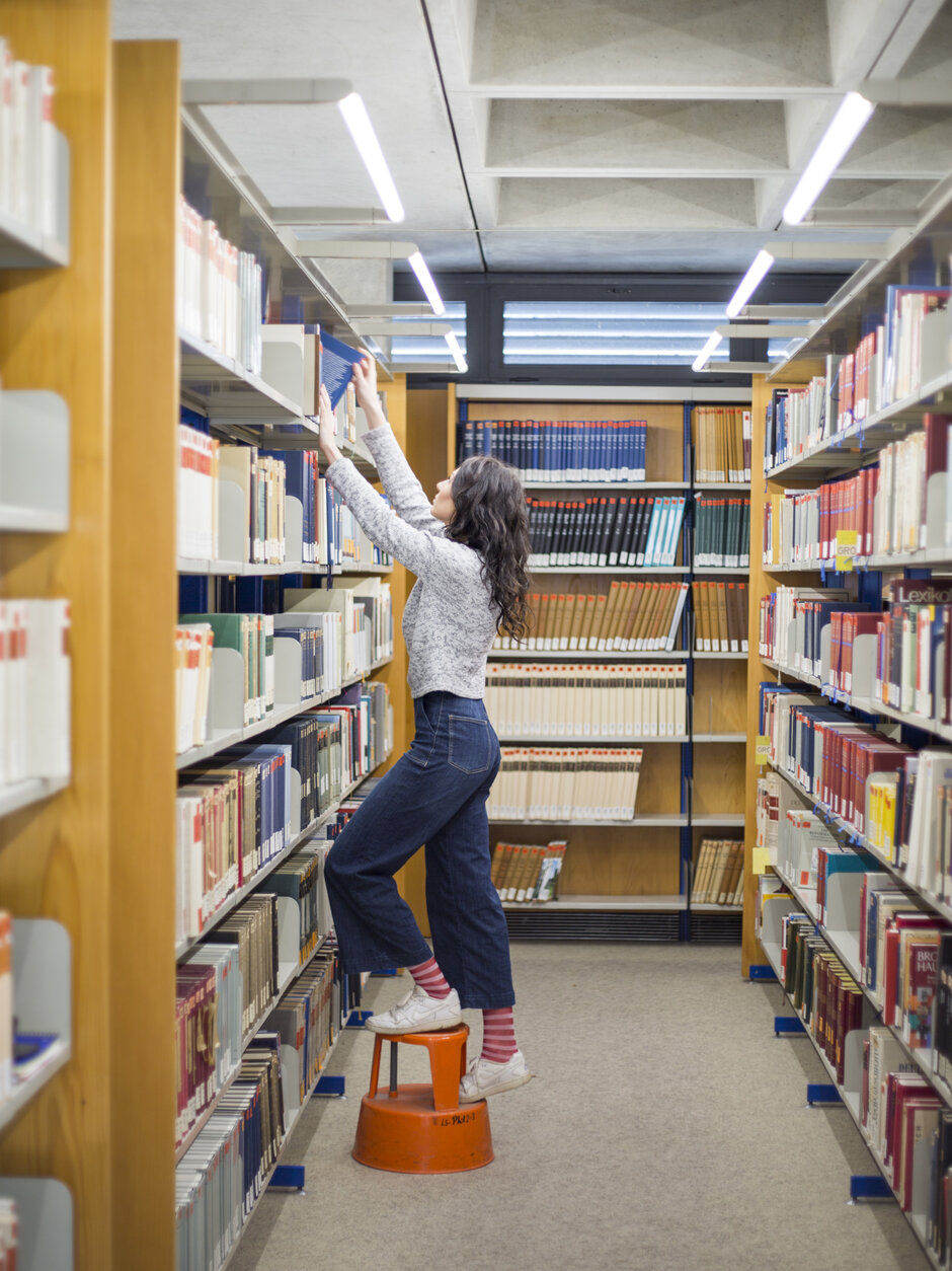 Eine junge Frau steht auf einem orangenen Schemel, um ein Buch aus dem obersten Regalfach eines Bücherregals zu holen.