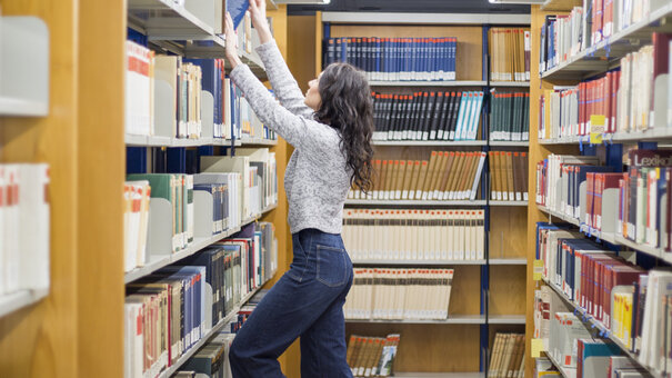 Eine junge Frau steht auf einem orangenen Schemel, um ein Buch aus dem obersten Regalfach eines Bücherregals zu holen.
