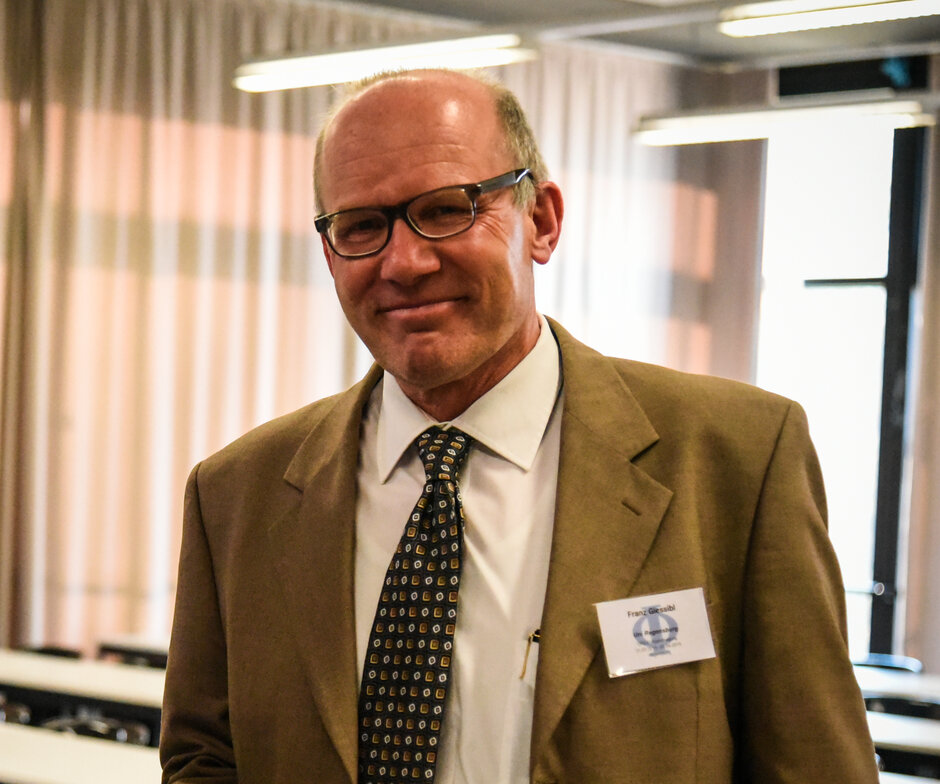 Ein ca. 60 Jahre alter Mann im braunen Anzug lächelt direkt in die Kamera.