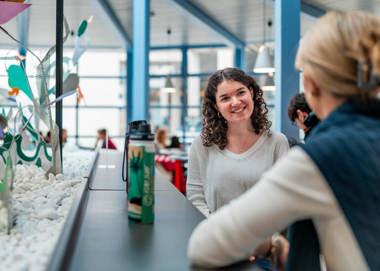 Zwei Frauen sitzen einander zugewandt an einem langen, thekenartigen Tisch in einem hellen Raum.