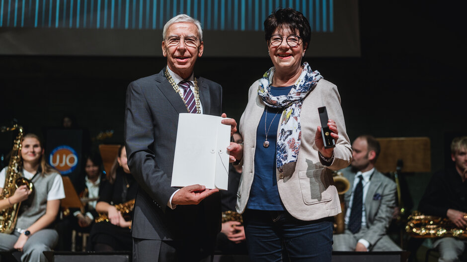Ein Herr in dunklem Anzug hält eine Urkundenmappe hoch. Neben ihm steht eine fröhliche Dame im hellen Blazer.