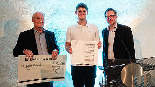 Drei Herren - die beiden außen in dunklen Sakkos, der mittlere in einem weißen Polo-Shirt - stehen nebeneinander. Der links Stehende hält einen Dekoscheck hoch, der mittlere eine aufgeschlagene Urkundenmappe.