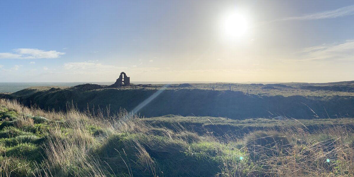 Küstenlandschaft, mit Gras bewachsen. Die Sonne steht oben am Himmel. Eine Ruine ist zu sehen. Im Hintergrund das Meer.