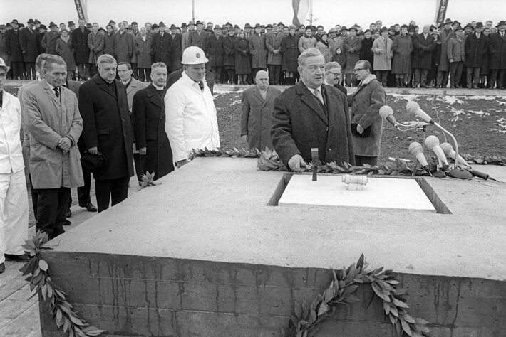 Das schwarz-weiß Foto zeigt Männer und Frauen in Wintermänteln, die sich um einen mit Girlanden umwundenen Betonsockel scharen. In der Bildmitte ist ein Mann zu sehen, der einen Hammer über eine in den Sockel eingelassene Kassette hält. (opens enlarged image)