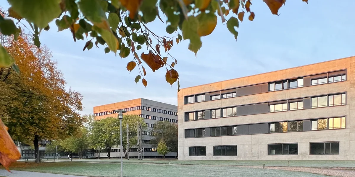 Zwei Hörsaalgebäude im Morgenlicht, umgeben von herbstlich gefärbten Bäumen. 
