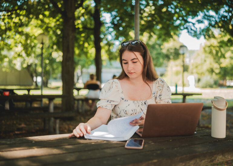 Eine Studentin sitzt mit Laptop und Studienunterlagen im Freisitz der Chemie-Cafete an der Universität Regensburg