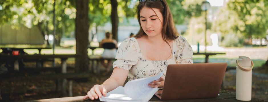 Eine Studentin sitzt mit Laptop und Studienunterlagen im Freisitz der Chemie-Cafete an der Universität Regensburg