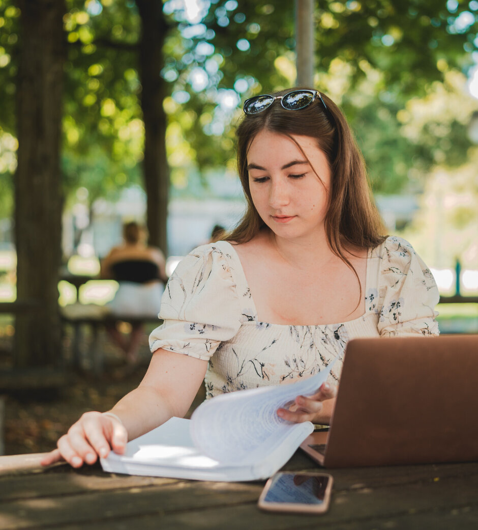 Eine Studentin sitzt mit Laptop und Studienunterlagen im Freisitz der Chemie-Cafete an der Universität Regensburg