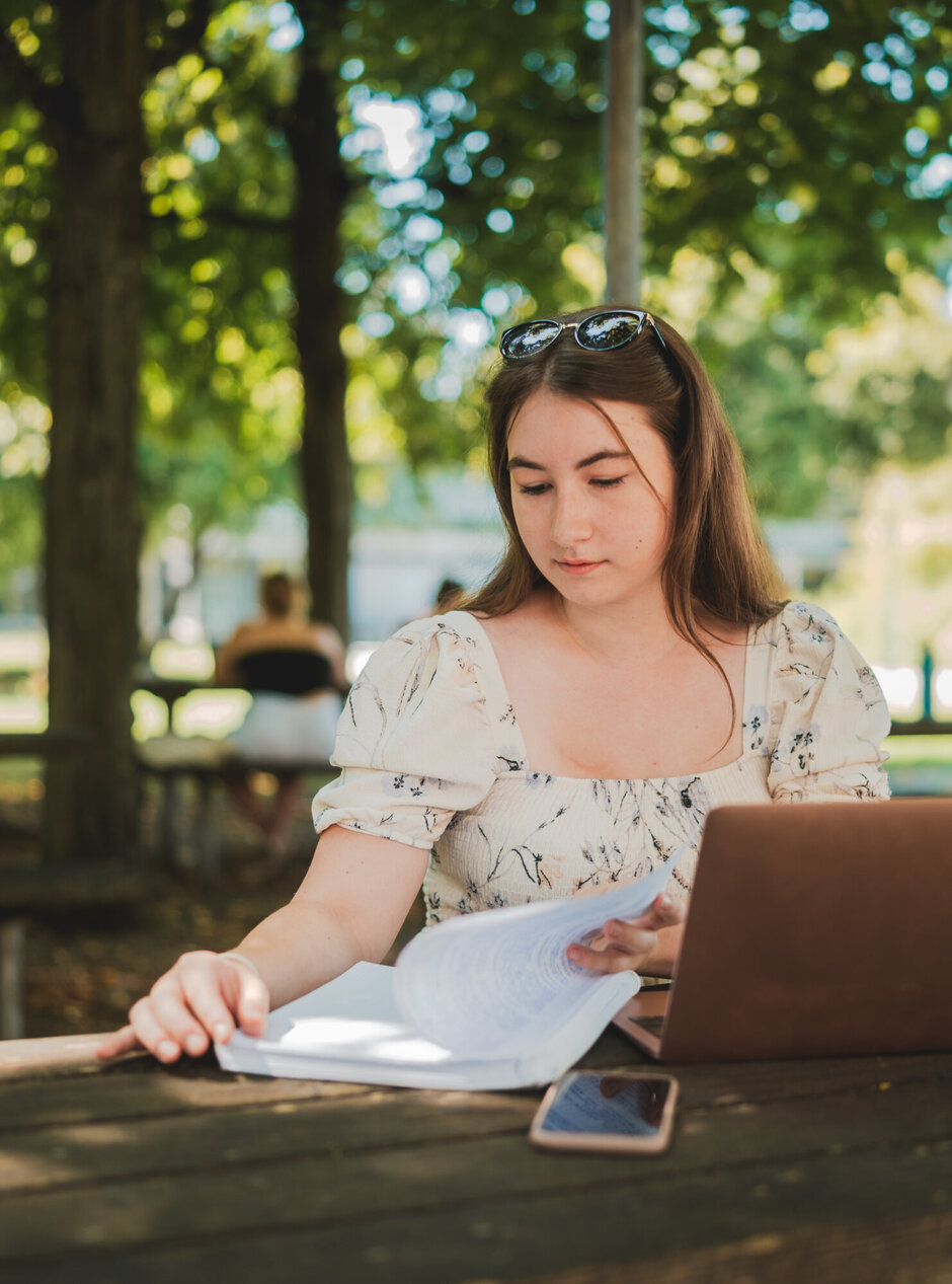 Eine Studentin sitzt mit Laptop und Studienunterlagen im Freisitz der Chemie-Cafete an der Universität Regensburg