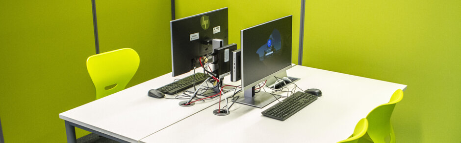 Zwei Schreibtische, die sich an der Längsseite direkt gegenüberstehen; auf jedem Schreibtisch ein Computerarbeitsplatz. Am Schreibtisch sind drei grüne Bürostühle zu sehen, die Schreibtische sind von grünen Trennwänden umgeben. 