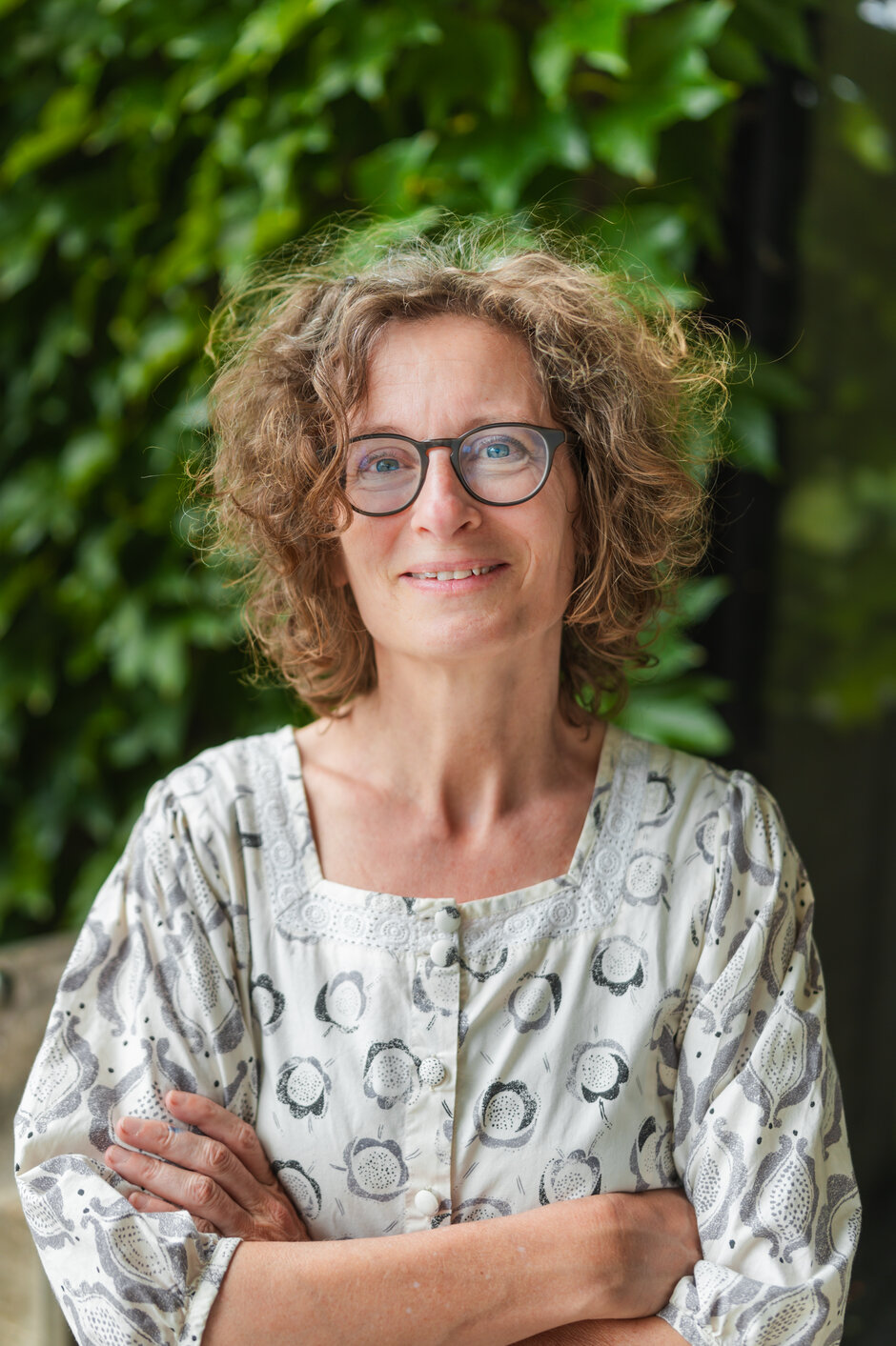 Eine freundliche Frau mit Brille und lockigem Haar steht vor einer Hecke und schaut den Betrachter an. Sie hat ihre Arme lässig vor dem Körper verschränkt.