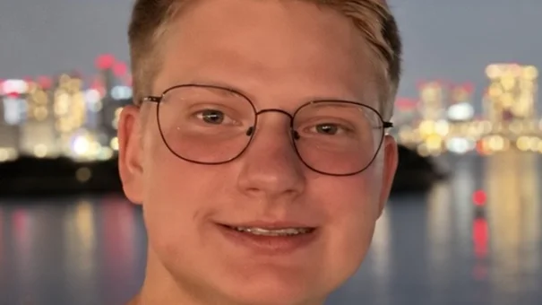 A portrait photograph of a young man with dark blond hair and round glasses, standing on the bank of a river at night, with illuminated buildings in the background.