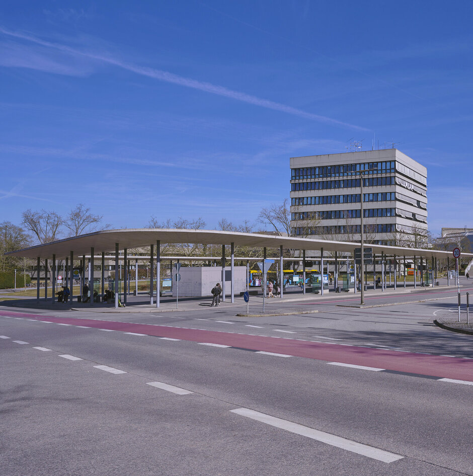 Eine große Bushaltestelle vor einem mehrstöckigen Gebäude in Sichtbetonbauweise.