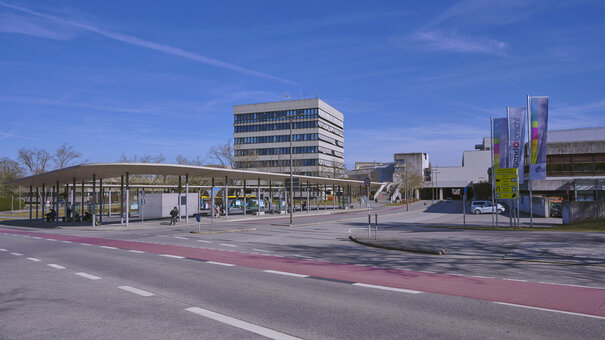 Eine große Bushaltestelle vor einem mehrstöckigen Gebäude in Sichtbetonbauweise.