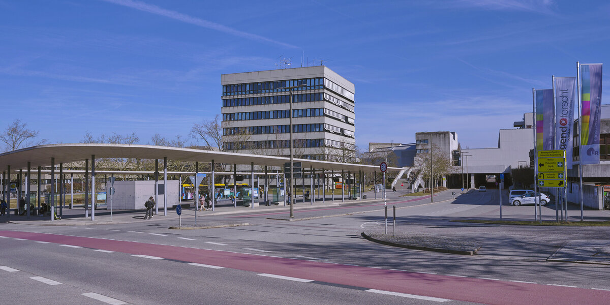 Eine große Bushaltestelle vor einem mehrstöckigen Gebäude in Sichtbetonbauweise.