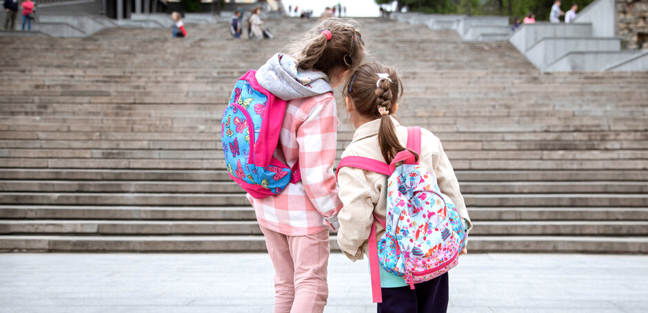 Zwei Kinder im Grundschulalter mit rosa-gemusterten Rücksäcken halten sich an der Hand und gehen auf eine Treppe zu.