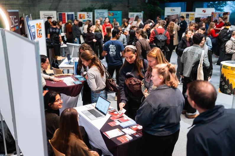 Viele Studierende informieren sich an Ständen im Foyer des Audimax der UR. An mehreren Tischen werden Informationsmaterialien ausgelegt und Gespräche geführt. (öffnet Vergrößerung des Bildes)