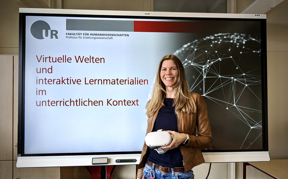 Eine Frau mit langen blonden Haaren und einer Lederjacke hält eine VR-Brille in der Hand. Sie steht vor einem großformatigen Bildschirm auf dem zu lesen steht "Virtuelle Welten und interaktive Lehrmaterialien im unterrichtlichen Kontext." (öffnet Vergrößerung des Bildes)