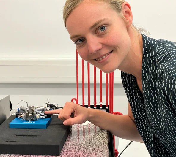 Eine junge Frau beugt sich lächelnd zu einem kleinformatigen elektronischen Gerät, auf das sie mit dem kleinen finger ihrer rechten Hand deutet.  (öffnet Vergrößerung des Bildes)