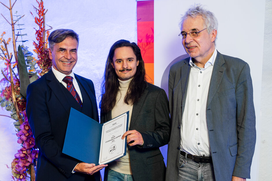 Drei Männer stehen vor einem Blumengesteck und einem Rollup mit farbigen Blockstreifen. Der jüngere, in der Mitte stehende hält eine Urkunde in die Kamera.